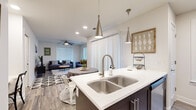 This one-bedroom, two-bathroom residence in Centerton, AR is thoughtfully designed to maximize every inch of its single-floor layout. The open-concept kitchen and living area serves as the heart of the home, featuring a large island with a double stainless steel sink, white quartz countertops, and dark espresso cabinetry extending to the ceiling for substantial storage. A gray tile backsplash complements a full suite of stainless steel appliances, including a refrigerator, range, and built-in microwave. Pendant lighting above the island anchors the space, which flows seamlessly into the living area. The living room benefits from two large windows that invite natural light, wood-style flooring, and a ceiling fan overhead. The bedroom offers plush carpeting, a ceiling fan, a dedicated window for natural light, and access to a closet. Both bathrooms feature tile surround tub-shower combinations and clean, neutral finishes. A connecting hallway links all rooms efficiently, contributing to the home's cohesive and functional layout throughout.