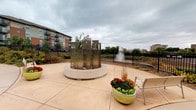 This contemporary residential building presents a polished and well-appointed common area designed with both function and aesthetic appeal in mind. The lobby entrance features a striking curved ceiling with warm gold and deep brown tones, complemented by stone-tile flooring that transitions into carpeted areas. Large windows flood the welcoming seating area with natural light, which is furnished with modern chairs and a sofa arranged around a bold red accent table — ideal for residents and guests alike. Stainless steel elevator doors are framed by dark tile surrounds, lending a sleek, upscale feel to the space. Adjacent to the elevators, a dedicated mailroom alcove houses an extensive array of individual mailboxes, providing convenient mail access for all residents. Curved hallways extend from the lobby, finished with patterned carpeting, recessed lighting, and dark wood paneling that maintains the building's refined design language throughout. Glimpses of additional amenity spaces, including what appears to be a fitness room, can be seen along the corridor. Outside, a paved courtyard serves as a tranquil retreat, centered around a sculptural stainless steel water feature set on a circular base. Colorful seasonal plantings and a nearby water view with a fountain visible beyond the perimeter fence complete this inviting outdoor environment.