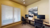 This single-floor business center is designed to provide a practical and focused workspace. The room is finished with warm yellow walls accented by crown molding, giving the space a polished yet approachable feel. Recessed and track lighting overhead ensures consistent, well-distributed illumination throughout the work area. A large window fitted with horizontal blinds allows for adjustable natural light, contributing to a bright and comfortable environment. The workstation area features a continuous granite-topped desk running along two walls, accommodating multiple computer monitors and ergonomic task chairs, making it well-suited for individual or concurrent use. A multifunction printer and scanner is conveniently positioned on the desk surface, supporting a range of document needs. A framed cityscape artwork adds a decorative touch to the otherwise functional space. Carpeted flooring helps reduce noise and adds a layer of comfort underfoot. Overall, the business center is thoughtfully outfitted to meet the everyday technology and administrative needs of its users.