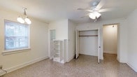 This single-floor space presents a clean, neutral aesthetic throughout, with bright white walls and trim that create an open, airy atmosphere. The bedroom is a standout feature, finished with plush wall-to-wall carpeting in a warm beige tone that adds comfort underfoot. Crown molding lines the upper walls, lending a subtle architectural detail that elevates the room's character. A ceiling fan with an integrated light fixture is centrally mounted, providing both air circulation and ambient lighting. Storage is well-addressed in the bedroom, with a full-width reach-in closet fitted with a hanging rod, offering ample space for clothing and accessories. A freestanding white louvered shelving unit positioned nearby provides additional open storage and organizational flexibility. Natural light filters softly into the space, contributing to the overall bright and welcoming feel. The layout is thoughtfully arranged, with clear sightlines to adjacent areas through open doorways, suggesting a connected and functional floor plan. The overall ambiance is understated and move-in ready, ideal for those who appreciate a simple, well-maintained living environment with practical storage solutions.