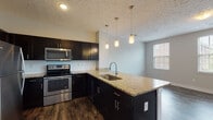 This well-appointed single-floor residence offers a thoughtfully designed open-concept layout that seamlessly connects the kitchen and living areas. The kitchen serves as the centerpiece of the space, showcasing dark espresso cabinetry, light granite countertops, and a full suite of stainless steel appliances including a refrigerator, electric range, and built-in microwave. A peninsula island with an undermount sink and elegant pendant lighting above adds both functionality and visual appeal. Rich, dark hardwood-style flooring runs throughout the kitchen and living areas, creating a warm and cohesive aesthetic. The spacious living area is bathed in natural light from a trio of windows, providing a bright and airy atmosphere. The bedroom features plush carpeting and direct access to a hallway leading to additional rooms. The bathrooms are finished with white vanities, light countertops, tile flooring, and chrome fixtures. The primary bathroom includes a walk-in shower with a sliding glass door, while a second full bathroom offers a matching vanity and mirror. Textured ceilings are present throughout, and neutral wall tones complement the overall modern and clean design of the space.