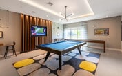 This dynamic single-floor amenity space is thoughtfully designed to encourage recreation, socializing, and casual work in equal measure. The layout flows seamlessly between distinct zones, each with its own character while maintaining a cohesive contemporary aesthetic.

The game room anchors one end of the space, highlighted by a full-size pool table with a vivid blue felt surface, set atop a boldly patterned area rug. A sculptural multi-arm pendant light hangs overhead within a coffered ceiling detail with integrated ambient lighting. Large windows with sheer curtains allow abundant natural light and views of the outdoors. A foosball table and additional game furnishings further enrich this entertainment zone.

Transitioning into the social lounge area, the flooring shifts to warm-toned hardwood, and the ceiling features striking wooden slat detailing alongside white structural columns that define the open corridor. Eclectic seating arrangements—including striped bar stools, houndstooth accent chairs, and marble-top tables—are distributed throughout, encouraging casual gatherings.

A dramatic full-wall mural with an urban, painterly motif serves as a bold focal point, while a built-in counter with bar stools offers a space for informal work or conversation. Smaller nook areas with wall-mounted desks and designer pendant lighting provide quieter spots for focused tasks. The overall ambiance is lively, modern, and welcoming.