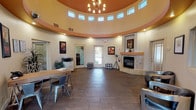 This single-floor space makes an immediate impression with its dramatic rotunda-style architecture. The circular main room draws the eye upward to a soaring domed ceiling accented in warm terracotta and cream tones, encircled by a ring of clerestory windows that flood the interior with abundant natural light. A classic chandelier hangs at the center of the dome, adding an elegant focal point to the open layout below.

The flooring throughout is a rich, dark wood-look tile that grounds the space and complements the warm color palette of the walls. A stone-surround fireplace anchors one side of the room, providing both a functional and decorative centerpiece that contributes to the inviting ambiance. Framed artwork is thoughtfully displayed on the curved walls, adding character and visual interest.

The space is furnished with a modest desk and metal chair, suggesting a functional reception or workspace area, while a lounge seating area is partially visible, indicating versatility in how the room is used. Decorative poufs add casual, flexible seating options. A glazed entry door provides a view to the exterior, further enhancing the sense of openness. Overall, the space combines architectural distinction with a warm, welcoming atmosphere suited for gathering, reception, or community use.