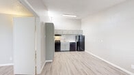 This single-floor unit offers a clean, functional layout with a cohesive design carried throughout each room. The kitchen serves as a standout feature, showcasing gray shaker-style cabinetry — including a full-height pantry cabinet for ample storage — paired with white subway tile backsplash and light countertops. Black appliances, including a gas range, dishwasher, and refrigerator, provide a modern contrast to the soft gray and white palette. Light wood-look plank flooring runs seamlessly throughout the space, adding warmth and continuity. A textured ceiling adds a subtle layer of character across the main areas. The living area is bright and open, benefiting from natural light through a window fitted with horizontal blinds, and is equipped with a wall-mounted HVAC unit for climate control. The bathroom is straightforward and functional, featuring a full-size tub and shower combo with white tile surround, a toilet, and a vanity with storage. The overall ambiance of the unit is clean and freshly updated, with a neutral color palette of whites and warm grays that creates an inviting, move-in-ready atmosphere throughout.
