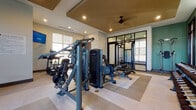 Located at 17014 Fairmount Way in Charlotte, NC, this single-floor fitness center is a comprehensively outfitted exercise facility designed to support a wide range of workout routines. The space is thoughtfully organized into distinct functional zones, each tailored to a specific type of training.

The main workout area features premium True-brand cardio equipment, including treadmills, ellipticals, and a stair climber, positioned beneath recessed lighting and wall-mounted flat-screen TVs for an engaging exercise experience. Natural light filters in through multiple windows fitted with roller shades, creating a bright and energizing atmosphere.

A dedicated strength-training zone houses a multi-station cable and weight machine, along with a rack of dumbbells, providing ample options for resistance training. A separate enclosed studio, accessible through sleek black-framed glass doors, is stocked with kettlebells, medicine balls, and functional training accessories — ideal for group classes or individual dynamic workouts.

Design details include a tray ceiling with warm gold tones accented by a ceiling fan, neutral-toned flooring with area rugs defining training zones, and sage green accent walls that lend a calm, modern aesthetic. Glass partitions and doors maintain an open, connected feel throughout the space, while an adjacent community lounge is visible through the entryway, adding to the facility's welcoming character.
