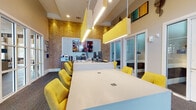 This single-story business center in Jacksonville, Florida, is designed to support a productive and professional environment through thoughtful layout and contemporary aesthetics. The space is defined by soaring ceilings that amplify its open, airy character, while large windows along one wall flood the interior with natural light and offer views of lush landscaping and palm trees outside.

The main workspace features a generously sized white communal table surrounded by vibrant yellow upholstered chairs, suspended above by modern linear pendant lights with wood accents. A long counter-height workstation with matching yellow seating lines one wall, providing additional workspaces beneath the high windows. A striking herringbone-patterned wood accent wall adds warmth and visual depth to the room.

Two glass-walled private offices with sliding doors provide enclosed, focused workspaces, each equipped with a desk and chair. A dedicated service station along one wall includes a coffee and beverage setup, a mini refrigerator, and a large wall-mounted display screen above. A printer station and reception desk with computer monitors complete the functional offerings. A comfortable lounge nook with neutral upholstered chairs and a round side table offers a relaxed setting for informal meetings or quiet work.