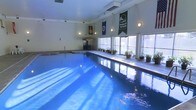 This single-level indoor pool facility offers a welcoming and functional aquatic environment. The centerpiece is a generously sized rectangular swimming pool with brilliantly clear blue water, ideal for both lap swimming and leisure. The pool deck surrounds the water with ample space for movement and relaxation, and is accented by a collection of potted plants and greenery that add a touch of life and color to the setting.

A standout feature of the space is a tiki-style bar area positioned along one side, complete with seating, adding a recreational and social dimension to the facility. Decorative banners hang from the walls, lending a spirited character to the room. Wall-mounted sconce lighting provides a warm, ambient glow throughout the interior.

Large windows line one wall of the pool area, inviting abundant natural light that reflects beautifully off the water's surface, enhancing the bright and airy atmosphere of the space. The light-colored walls and ceiling contribute to the open, clean aesthetic of the room. A ceiling-mounted HVAC unit ensures climate comfort year-round, making this an all-season aquatic destination. Overall, the space strikes a balance between practical athletic use and inviting recreational appeal.