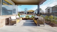 This covered outdoor lounge area serves as a poolside retreat within a residential community, offering a well-appointed space for relaxation and socializing. The structure features a solid overhead covering supported by columns with brick detailing at the base, providing shade while maintaining an open, airy feel with direct sightlines to the swimming pool and surrounding landscape.

The lounge is furnished with wicker and rattan-style seating accented in yellow and white cushions, including both individual chairs and a sectional sofa arrangement. A wall-mounted flat-screen television is installed under the covered section, creating an entertainment-ready outdoor environment ideal for gatherings. The polished concrete flooring runs throughout the covered area, transitioning seamlessly to the pool deck beyond.

Posted pool rules and signage are prominently displayed, noting dawn-to-dusk operating hours. The pool area beyond features additional seating, yellow market umbrellas, and mature plantings that define the space. Views from the lounge extend to multi-story residential buildings surrounded by trees, providing a sense of community scale. Natural light fills the open sides of the structure, enhancing the inviting ambiance of this shared amenity space.