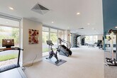 Located at 526 Hartford Tpke in Shrewsbury, MA, this fitness facility offers a comprehensive, single-floor layout designed to support a wide range of workout activities. The space is branded as "The Elmwood" and is organized into distinct zones, each serving a specific fitness purpose.

The main exercise room is generously proportioned, featuring light-toned carpeting, recessed lighting, and floor-to-ceiling windows that flood the space with natural light and provide views of the surrounding greenery. This area is equipped with cardio machines including treadmills, stair climbers, and stationary bikes, as well as strength training equipment such as cable machines, a multi-functional rack, kettlebell storage, and TRX suspension systems. Large wall-mounted mirrors enhance the sense of space and support proper form during workouts.

A separate studio room features bold teal walls, a drop ceiling with recessed lighting, full-length mirrors, and carpeted flooring — ideal for yoga, stretching, or group fitness classes. An additional area with a teal and orange color scheme serves as a reception or transitional zone, complete with built-in cabinetry and informational displays.

Throughout the facility, large windows maintain a bright, open atmosphere, while the modern design and variety of equipment make this a well-rounded and functional fitness destination.