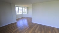 This single-floor space offers a clean, contemporary design characterized by an open, airy layout and a neutral color palette throughout. The main living area is anchored by rich, dark hardwood-style flooring that contrasts beautifully with the crisp white walls, creating a warm yet modern aesthetic. A standout feature is the large corner window arrangement, which floods the space with abundant natural light and frames elevated views of the surrounding urban landscape, including greenery and city buildings beyond. A pendant light fixture hangs overhead, adding a subtle decorative touch that complements the understated elegance of the room. The generous, open floor plan provides a versatile canvas, allowing for a variety of furniture arrangements and uses. The smooth, well-maintained walls and clean baseboards reflect a space that has been thoughtfully maintained or freshly updated. The overall ambiance is bright, welcoming, and move-in ready, with the interplay of natural light, quality flooring, and urban vistas contributing to a sense of comfortable, modern living. The single-floor layout ensures easy accessibility and a cohesive flow throughout the entire space.