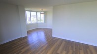 This single-floor space offers a clean, contemporary design characterized by an open, airy layout and a neutral color palette throughout. The main living area is anchored by rich, dark hardwood-style flooring that contrasts beautifully with the crisp white walls, creating a warm yet modern aesthetic. A standout feature is the large corner window arrangement, which floods the space with abundant natural light and frames elevated views of the surrounding urban landscape, including greenery and city buildings beyond. A pendant light fixture hangs overhead, adding a subtle decorative touch that complements the understated elegance of the room. The generous, open floor plan provides a versatile canvas, allowing for a variety of furniture arrangements and uses. The smooth, well-maintained walls and clean baseboards reflect a space that has been thoughtfully maintained or freshly updated. The overall ambiance is bright, welcoming, and move-in ready, with the interplay of natural light, quality flooring, and urban vistas contributing to a sense of comfortable, modern living. The single-floor layout ensures easy accessibility and a cohesive flow throughout the entire space.