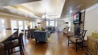 This thoughtfully designed single-floor space makes a strong impression with its open, airy layout and refined architectural details. The main living area is anchored by a striking tray ceiling with a coffered center section adorned with an elegant chandelier, adding a sense of grandeur and height to the room. Rich, warm hardwood flooring runs throughout, providing a cohesive and inviting foundation for the space.

Natural light pours in generously through multiple windows positioned along the walls, creating a bright and welcoming ambiance throughout the day. The living area is generously proportioned, offering ample room for multiple seating arrangements, including plush sofas and accent chairs in neutral and muted tones accented with pops of yellow.

A classic brick fireplace anchors one end of the room, lending a cozy, traditional character, while recessed lighting and decorative floor lamps complement the natural light. The open floor plan allows for seamless flow between the living and dining areas, making the space ideal for both relaxed everyday living and entertaining. Crown molding and carefully considered finishes throughout reflect a commitment to quality and attention to detail, resulting in a space that is both functional and elegant.