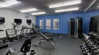This fitness center is a single-level space designed to provide a comprehensive exercise environment. The room is defined by clean white walls accented by a bold blue feature wall, creating an energizing atmosphere conducive to physical activity. Overhead, rows of fluorescent strip lighting ensure the space is brightly and evenly illuminated throughout.

The flooring consists of durable rubber gym matting, well-suited for heavy equipment and high-impact activity. Natural light filters in through multiple windows along the blue accent wall, fitted with horizontal blinds for adjustable privacy and glare control.

The cardio section is equipped with treadmills and an elliptical machine, offering options for both low-impact and higher-intensity aerobic workouts. A wall-mounted flat-screen television is positioned for viewing during cardio sessions. The strength training area includes an adjustable weight bench and a multi-function cable machine, providing versatility for a range of resistance exercises.

The layout is thoughtfully organized to allow comfortable movement between equipment, making the space functional for multiple users at once. Overall, the fitness center presents a practical, well-maintained environment suited for residents or members seeking convenient access to essential workout amenities.