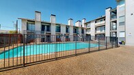 This residential complex is centered around a well-maintained outdoor swimming pool, which serves as the focal point of the communal outdoor area. The pool is enclosed by a sturdy metal safety fence and is framed by a gravel-and-aggregate courtyard that provides a durable, low-maintenance surface throughout the common grounds. Surrounding the pool on multiple sides are multi-story residential buildings characterized by a stucco exterior, distinctive protruding chimney structures, and private balconies accessible from upper-level units. Exterior metal staircases connect the ground level to the upper floors, adding a functional and industrial aesthetic to the overall design. A separate single-story ancillary building is also present on the property, featuring blue-tinted grid windows and a stucco facade, which appears to serve a utility or laundry function. Horizontal wood-slat fencing panels add a warm, contemporary design element and define portions of the perimeter. The entire complex is set beneath open skies, offering an abundance of natural light throughout the outdoor spaces. The architectural style reflects a mid-century multi-unit residential design, combining practical construction with communal amenities suited for shared residential living.