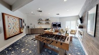 This single-floor game and lounge room is designed as a dynamic social hub, blending entertainment and relaxation in a well-appointed, open layout. The space is anchored by a bold navy and gold starburst-patterned carpet that sets an energetic tone throughout. Recessed lighting illuminates the room evenly, complementing the natural light that enters through exterior-facing doors and windows.

A standout feature is the textured wood-plank accent wall, which houses a sleek linear fireplace and a wall-mounted flat-screen television, flanked by curated artwork. Opposite this, a full-size foosball table serves as the centerpiece of the room, while two classic arcade machines provide additional gaming entertainment near the windowed wall.

A built-in wet bar area along one wall includes lower cabinetry with a countertop, a sink, and a microwave, complemented by open floating shelves styled with decorative accents. Comfortable lounge seating, including a tufted leather sofa, patterned accent chairs, and a round wood coffee table, creates an inviting gathering area. A large framed wall-mounted Scrabble board adds a playful, interactive touch. The space connects to an adjacent room through glass-paneled black doors.