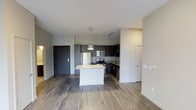 This single-floor space showcases a thoughtfully designed open-concept layout that seamlessly connects the kitchen and living areas. The kitchen is a standout feature, anchored by a central island topped with a light granite surface and accented by a contemporary pendant light overhead. Dark-toned cabinetry lines the kitchen walls, providing a sleek contrast to the lighter countertops and neutral-colored walls. A full suite of stainless steel appliances — including a refrigerator, range, microwave, and dishwasher — adds a polished, modern touch. Track lighting runs along the ceiling, ensuring the kitchen is well-lit and functional. The flooring throughout consists of wood-style planks in a warm, medium-toned finish, lending continuity and warmth across the open space. The living area benefits from the open layout, creating a generous and airy feel. Walls are finished in a clean, neutral palette, complementing the modern aesthetic. A dark entry door provides a striking contrast near the entrance. The overall ambiance is contemporary and clean, making the space feel both functional and inviting.