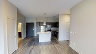 This single-floor space showcases a thoughtfully designed open-concept layout that seamlessly connects the kitchen and living areas. The kitchen is a standout feature, anchored by a central island topped with a light granite surface and accented by a contemporary pendant light overhead. Dark-toned cabinetry lines the kitchen walls, providing a sleek contrast to the lighter countertops and neutral-colored walls. A full suite of stainless steel appliances — including a refrigerator, range, microwave, and dishwasher — adds a polished, modern touch. Track lighting runs along the ceiling, ensuring the kitchen is well-lit and functional. The flooring throughout consists of wood-style planks in a warm, medium-toned finish, lending continuity and warmth across the open space. The living area benefits from the open layout, creating a generous and airy feel. Walls are finished in a clean, neutral palette, complementing the modern aesthetic. A dark entry door provides a striking contrast near the entrance. The overall ambiance is contemporary and clean, making the space feel both functional and inviting.