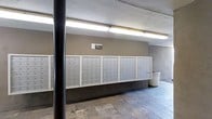 The mail center is a functional, communal space designed to efficiently serve a large number of occupants. The area is defined by an expansive wall-mounted bank of individual aluminum mailboxes, organized into multiple columns and rows, accommodating a significant number of users. A clearly posted "Mail Center" sign identifies the space, making it easy to locate. The floor is finished with stone or slate-style tiles in natural, earthy tones, adding a degree of texture and durability suited to a high-traffic area. The walls are finished in a smooth taupe/grey plaster, contributing to a neutral and utilitarian aesthetic. Overhead, fluorescent lighting provides bright, even illumination throughout the space. A structural support column is visible in the foreground, indicating a robust construction. A waste bin is conveniently positioned near the mailboxes for easy disposal. Overall, the space is practical and straightforward in design, prioritizing accessibility and functionality for a large community of users.