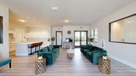 Located at 3681 Michigan Ave in Fort Myers, Florida, this single-floor community space offers a well-appointed and thoughtfully designed layout ideal for gathering and relaxation. The open-plan interior is finished with light wood-look flooring that runs seamlessly throughout, complementing the crisp white walls and creating a bright, airy atmosphere.

The expansive lounge area serves as the heart of the space, furnished with plush emerald-green velvet sofas arranged around a contemporary oval coffee table. Multiple windows along the perimeter flood the room with natural light, while glass double doors provide direct access to the outdoor community grounds and further enhance the sense of openness. A wall-mounted television and additional seating areas make this a versatile space for both casual socializing and organized events. A pass-through window connects the lounge to an adjacent office or workstation area, adding functional flexibility.

The kitchen is clean and modern, featuring an extensive run of white shaker-style cabinetry with brushed nickel hardware, expansive white countertops, and a stainless steel refrigerator. A large central island with bar-height seating accommodates multiple guests, making it well-suited for community gatherings. The overall design of the space strikes a balance between contemporary style and practical functionality, offering residents a welcoming and comfortable shared environment.