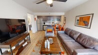 Located at 207 E Mountcastle Dr in Johnson City, Tennessee, this single-floor unit offers a thoughtfully designed layout with a cohesive modern aesthetic accented by bold orange décor throughout. The open-concept living area features light hardwood flooring, a ceiling fan, and a large front-facing window that allows natural light to fill the space. A leather sofa, upholstered accent chair, and a rustic wood coffee table anchor the living room, while a flat-screen TV on an industrial-style media console adds functionality. The living area flows seamlessly into the kitchen, which is outfitted with dark wood cabinetry, a subway tile backsplash, light quartz countertops, and a full suite of stainless steel appliances including a refrigerator, range, microwave, and dishwasher. A small dining area with a bistro table and orange chairs sits adjacent to the kitchen. The bedroom is a comfortable retreat featuring a queen bed with a rustic wooden headboard, a dresser with mirror, ceiling fan, a closet, and artwork that ties into the unit's vibrant color palette. The full bathroom includes a tub-shower combination with white tile surround, hardwood-style flooring, and dark vanity cabinetry with a white countertop.