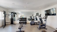 This well-appointed fitness facility comprises two dedicated exercise rooms, together offering a comprehensive workout environment with a clean, modern design. Crisp white walls, recessed lighting, and smooth concrete-style flooring create a bright, professional atmosphere throughout the space.

The first exercise room is oriented around strength and cardio training, featuring a full rack of dumbbells, elliptical trainers, a stationary bike, and an Assault air bike. Floor-to-ceiling mirrors line one wall, visually expanding the room and allowing users to monitor their form. A wall-mounted flat-screen television adds to the functionality of the space.

The second exercise room is dedicated to cardio equipment, housing two commercial-grade treadmills and a rowing machine, also accompanied by a wall-mounted television. This room benefits from direct access to an outdoor area through a glazed door, allowing natural light to filter in. Windows throughout both rooms frame scenic mountain views, creating an energizing backdrop for workouts.

Additional equipment visible across the facility includes a cable machine, a back extension bench, a spin bike, and a weight bench, offering a well-rounded selection for varied training needs. The overall design is functional and uncluttered, prioritizing ease of movement and a motivating environment for fitness.