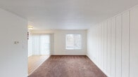 This single-floor unit in Indianapolis, IN offers a straightforward and functional layout spread across several distinct areas. The living room serves as the central gathering space, featuring plush carpeting, a distinctive vertically paneled accent wall, and generous natural light drawn in through a large window and a sliding glass door dressed with vertical blinds. The transition from carpet to hardwood-style flooring marks the entry into the dining and kitchen area, which is connected via a half-wall pass-through, creating an open sightline between the two spaces. The galley-style kitchen is well-appointed with white cabinetry offering ample storage, dark-toned laminate countertops, a double-basin sink, and white appliances including an electric range and refrigerator. The bedrooms continue the carpeted flooring and maintain a clean, neutral palette with bright white walls and windows that invite natural light. One bedroom features access to a closet space. The bathroom is finished in crisp white tones, complete with a vanity, mirror with Hollywood-style lighting, and hardwood-style flooring. Throughout the unit, white walls and consistent flooring materials contribute to a cohesive, light, and airy atmosphere.