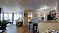 This single-floor residence showcases an open-concept design that seamlessly connects the kitchen, dining, and living areas into one cohesive and functional space. Rich hardwood flooring runs throughout, anchoring the warm, neutral color palette of beige and tan tones that define the interior.

The kitchen is equipped with a peninsula-style breakfast bar featuring a granite or stone countertop in a cool blue-grey tone, complemented by bar stools that invite casual dining. Track lighting overhead provides focused illumination across the kitchen and adjoining areas. A stainless steel sink and ample counter space add to the kitchen's practicality.

The living area is spacious and bright, benefiting from natural light filtering through large windows dressed with curtains. A comfortable sofa, side table, and decorative lamp contribute to a welcoming atmosphere. An accent wall in soft blue adds a subtle pop of color to the living space.

A doorway visible from the main living area leads to a bedroom, decorated with vibrant orange and patterned textiles, providing a striking contrast to the calm tones of the main living space. A textured popcorn ceiling spans the entire unit, and tasteful wall art and potted plants add decorative character throughout the home.