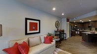 This single-floor space offers a thoughtfully designed open-concept layout that seamlessly connects the living area to the kitchen. The living room is warm and inviting, anchored by a cream-toned sofa accented with bold red throw pillows that complement the decorative framed artwork on the wall. Recessed lighting throughout the ceiling provides consistent, modern illumination across the space. Hardwood-style flooring runs throughout, adding warmth and continuity between rooms. A small bistro-style dining area with bar stools serves as a transitional zone between the living and kitchen spaces, ideal for casual dining or entertaining. The kitchen, visible in the background, features stainless steel appliances including a refrigerator and built-in microwave, set against dark cabinetry that provides a rich contrast to the lighter tones of the rest of the space. Decorative touches such as a large vintage-style wall clock and an "EAT" sign above the kitchen entrance add character and personality. The neutral gray walls serve as a versatile backdrop that ties together the warm and bold design elements. Overall, the space presents a balanced blend of comfort, functionality, and style.