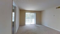 This single-floor residential space offers a clean and functional layout designed for comfortable everyday living. The main living area is a standout feature, characterized by neutral beige walls, plush wall-to-wall carpeting, and white baseboards that lend the room a tidy, cohesive aesthetic. A large sliding glass door dominates the far wall, fitted with vertical blinds that allow for adjustable light control. The door opens to a private balcony enclosed by white railings, beyond which lush tree foliage provides a pleasant natural backdrop and a sense of privacy. Natural light filters generously into the living space through both the sliding door and an additional window visible on the adjacent wall, keeping the interior feeling bright and airy throughout the day. The smooth, white ceiling contributes to an open and uncluttered atmosphere. The overall ambiance of the space is calm and understated, with a neutral color palette that offers a versatile canvas for a variety of interior styles. The connection to the outdoors through the balcony adds a welcoming dimension, making the living area feel extended and well-ventilated. This space is well-suited for those seeking a straightforward, low-maintenance living environment with a pleasant outdoor connection.