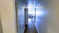 This single-floor space makes a strong first impression through its well-appointed hallway, which sets the tone for the interior's modern aesthetic. The corridor is defined by smooth grey-blue walls that create a calm, cohesive color palette throughout. Dark hardwood flooring runs the length of the hallway, grounding the space with warmth and contrast against the lighter wall tones. A panel ceiling adds a clean, finished look overhead, while a pendant light fixture provides focused illumination. The hallway is thoughtfully styled with curated wall art — notably a large blue-and-white textured piece that adds visual interest and an artistic sensibility to the passage. A door along the corridor provides access to an adjacent room. At the far end of the hallway, the space opens into a brighter, naturally lit living area, where glimpses of seating, a work surface, and additional artwork suggest a well-organized and stylishly furnished interior. The overall ambiance is contemporary and refined, with a consistent design language of cool tones, clean lines, and intentional decor choices that carry through from entry to living space.