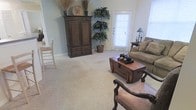This single-floor space offers a comfortable and classically styled interior that blends traditional design elements with a warm, welcoming ambiance. The living area is anchored by plush, light-toned wall-to-wall carpeting that extends throughout, lending a soft underfoot feel and a sense of cohesion to the space. Walls in a muted, calming blue-gray hue complement the rich, dark wood tones of the furnishings, creating a balanced and inviting atmosphere. A stately wooden armoire serves as a prominent focal point, topped with decorative accents and flanked by a lush potted tree that adds a natural, organic element to the room. A rustic wooden trunk-style coffee table anchors the seating area, paired with a plush neutral-toned sofa and an elegantly carved accent chair upholstered in warm leather tones. Natural light filters generously into the space through a full-height exterior door with sidelights and blinds, illuminating the room and enhancing its open, airy feel. A classic table lamp on a dark wood side table adds a layered lighting option for the evenings. The overall design aesthetic is traditional and well-appointed, offering a space that feels both functional and thoughtfully curated.