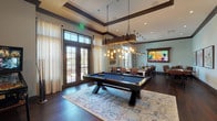 This single-floor amenity space is thoughtfully designed for recreation and socializing, combining refined finishes with a welcoming, lodge-inspired atmosphere. Rich dark hardwood flooring runs throughout, grounded by tray ceilings with dark wood crown molding that add architectural depth and visual interest to each area.

The main recreation zone centers on a full-sized pool table topped in blue felt, positioned beneath a striking rustic chandelier made from reclaimed wood and exposed Edison bulbs. Adjacent to it, a foosball table and wall-mounted flat-screen television framed in weathered wood offer additional entertainment options.

A long communal bar-height table with leather-upholstered chairs provides ample seating beneath modern pendant lights, while a built-in kitchenette features white cabinetry, quartz countertops, a deep sink, a beverage refrigerator, and a coffee station—all illuminated by oversized pendant fixtures.

A transitional lounge area features a pair of classic rocking chairs beside curated wall art, connecting seamlessly to adjoining rooms through glass-paned interior doors. Floor-to-ceiling windows and French doors bathe the space in natural light, offering views of palm trees and manicured outdoor grounds, enhancing the relaxed yet polished ambiance throughout.