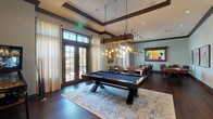 This single-floor amenity space is thoughtfully designed for recreation and socializing, combining refined finishes with a welcoming, lodge-inspired atmosphere. Rich dark hardwood flooring runs throughout, grounded by tray ceilings with dark wood crown molding that add architectural depth and visual interest to each area.

The main recreation zone centers on a full-sized pool table topped in blue felt, positioned beneath a striking rustic chandelier made from reclaimed wood and exposed Edison bulbs. Adjacent to it, a foosball table and wall-mounted flat-screen television framed in weathered wood offer additional entertainment options.

A long communal bar-height table with leather-upholstered chairs provides ample seating beneath modern pendant lights, while a built-in kitchenette features white cabinetry, quartz countertops, a deep sink, a beverage refrigerator, and a coffee station—all illuminated by oversized pendant fixtures.

A transitional lounge area features a pair of classic rocking chairs beside curated wall art, connecting seamlessly to adjoining rooms through glass-paned interior doors. Floor-to-ceiling windows and French doors bathe the space in natural light, offering views of palm trees and manicured outdoor grounds, enhancing the relaxed yet polished ambiance throughout.