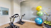 Located at 4045 George Busbee Pkwy NW in Kennesaw, Georgia, this dedicated exercise room offers a thoughtfully organized fitness space within a compact footprint. The room features light hardwood-style flooring and soft sage-green walls accented by crisp white crown molding and baseboards, creating a clean, energizing atmosphere.

The space is equipped with multiple stationary cycling bikes positioned on protective floor mats, providing a focused cardio area. A well-stocked equipment rack along one wall holds an assortment of stability balls in varying sizes, medicine balls, resistance bands, and foam rollers, offering versatile workout options. A plyo box adds functional training capability to the room's offerings.

Natural light filters in through large windows fitted with white plantation shutters, brightening the interior and offering views of the surrounding outdoor area. A wall-mounted flat-screen television provides entertainment during workouts. A full-length mirror enhances the sense of space while allowing users to monitor their form. The notably high ceiling height contributes to an open, airy feel despite the room's intimate scale. A glass-panel door connects the space to an adjacent fitness area, extending the overall exercise options available.