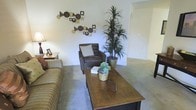 This single-floor residence offers a welcoming living area designed for everyday comfort and functionality. The main living room features plush wall-to-wall carpeting in a neutral tone, creating a warm and inviting atmosphere. The space is furnished with a striped sofa, a dark upholstered armchair with a coordinating accent pillow, and a solid wood rectangular coffee table that serves as a central anchor for the seating arrangement. Decorative touches include an eclectic geometric wall art arrangement featuring circular and angular pieces in earthy tones, adding visual interest to the light-colored walls. A tall, lush indoor plant in a woven basket brings a natural element into the room, complementing the smaller succulent centerpiece on the coffee table. An arched doorway is visible in the background, hinting at additional rooms beyond the living area. Natural and artificial lighting work together to keep the space bright and open. The overall ambiance is casual and livable, with a cohesive earthy color palette of browns, tans, and greens throughout the furnishings and décor.