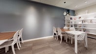 This thoughtfully designed library offers a refined and functional environment spread across a single floor. The space is organized into multiple seating areas, each furnished with white rectangular tables and contemporary chairs featuring elegant lattice and ghost-chair designs. The flooring throughout consists of neutral-toned carpet tiles that add warmth and comfort underfoot.

A defining feature of the library is its striking accent wall finished in a deep slate blue, which contrasts beautifully against the lighter walls and white furnishings. White open shelving units line the walls, displaying a curated collection of books, decorative vases, and accent pieces, adding both function and visual interest. A wooden sideboard with distinctive oval cutout detailing serves as a stylish storage piece along one wall.

Lighting is provided by a combination of recessed ceiling fixtures and pendant lights that cast a warm, even glow throughout the space. Sheer white floor-to-ceiling drapes line one wall, softening the room's atmosphere and contributing to its bright, airy feel. A computer workstation tucked into one corner provides a dedicated area for digital research. The drop ceiling with a grid tile system adds a clean, professional finish. Overall, the library exudes a modern yet welcoming character suited for reading, study, or quiet collaboration.