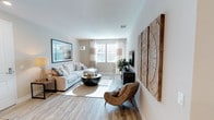 This single-floor space showcases a thoughtfully designed living area that balances contemporary style with comfortable functionality. The living room serves as the central gathering space, featuring light hardwood-style flooring that runs throughout and contributes to the room's warm, cohesive aesthetic.

Natural light flows in through a large window dressed with sheer white and neutral-toned curtains, brightening the space and highlighting the soft, neutral color palette of white and warm gray tones on the walls. The living area is well-proportioned, comfortably accommodating a plush, light-gray sectional sofa paired with a round, dark coffee table and a woven accent chair that adds a natural, organic texture to the room.

A standout decorative element is the large wooden wall art featuring circular grain patterns, adding a rustic yet refined focal point to the space. A wall-mounted flat-screen television is positioned opposite the seating area atop a sleek, dark media console. Additional framed artwork and a floor lamp enhance the layered, curated feel of the interior. Overall, the space presents a clean, modern ambiance with inviting warmth.