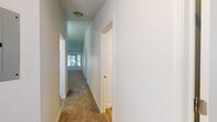 This single-story space is thoughtfully laid out, connecting its rooms through a carpeted hallway lined with textured walls and flush-mount ceiling lighting. The hallway leads toward a naturally lit area at its end, with doors branching off to various rooms along the way.

The kitchen is a functional highlight, showcasing white shaker-style cabinetry, light stone-look countertops, and a full suite of stainless steel appliances including a refrigerator, electric range with over-the-range microwave, and dishwasher. Wood-look plank flooring grounds the space with a warm, clean aesthetic.

The living and dining areas feature plush carpeting underfoot. The dining area is distinguished by a vaulted ceiling and an elegant chandelier, lending a sense of height and refinement. A window provides natural light and views of surrounding greenery.

The bedrooms are carpeted and well-proportioned, each featuring windows that allow natural light and outdoor views of mature trees. One bedroom includes a closet door, adding practical storage.

The bathroom features wood-look plank flooring, white cabinetry with stone-look countertops, and clean, bright finishes throughout. The overall ambiance of the space is neutral, bright, and move-in ready.