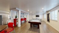 This single-floor amenity space is thoughtfully designed to serve as a multifunctional gathering and recreation area. The open-plan layout is anchored by warm wood-look flooring throughout, clean white walls with gray accents, and recessed lighting that keeps the space bright and welcoming.

The recreation zone dominates the main area, featuring a full-size pool table as its centerpiece, accompanied by a ping-pong table and a foosball table. Multiple wall-mounted flat-screen TVs are positioned for comfortable viewing from various angles, while bold red bar-height chairs and tables provide casual seating. Structural white columns define the space without obstructing sightlines, and a bar-height counter with red seating along one wall creates an ideal lounge and social hub.

Adjacent to the recreation area, a galley-style kitchen is fully outfitted with crisp white shaker cabinetry, granite-patterned countertops lining both sides, a stainless steel sink, and full-size appliances including a refrigerator and dishwasher. Recessed lighting and hardwood-style flooring continue seamlessly into the kitchen, maintaining a cohesive aesthetic. Natural light filters in through windows, adding to the clean, modern ambiance. This space is ideally suited as a community amenity room for entertaining and social gatherings.