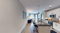 This single-floor residence on National Drive in Raleigh, NC, presents a thoughtfully designed open-concept layout with contemporary finishes throughout. The kitchen serves as a central hub, featuring white cabinetry extending to the ceiling, stainless steel appliances including a French door refrigerator and gas range, a subway tile backsplash, and a large island with a quartz countertop, undermount sink, and black pendant lighting overhead. Bar-height seating along the island connects the kitchen seamlessly to the living area, where light wood-style flooring continues underfoot and large windows draw in natural light. The living space offers a comfortable, well-scaled area anchored by a teal tufted sofa and vibrant wall art. The bedroom is a calm retreat with neutral tones, soft carpet, a ceiling fan, large windows with roller shades, and ample room for full furnishings. Adjacent, the bathroom features large-format marble-look tile in the shower, a floating vanity with a backlit mirror, and direct access to a walk-in closet with built-in shelving and hanging organization. Gray-trimmed interior doors and recessed lighting tie the space together with a clean, modern aesthetic.