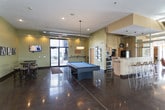 This single-floor amenity space is designed for social and recreational use, combining a comfortable lounge area with entertainment features. The centerpiece of the room is a full-size pool table with a vibrant blue felt surface, positioned beneath a modern drum pendant light that draws the eye and anchors the space. Polished concrete floors reflect the abundant natural light streaming in through multiple large windows, creating a bright and inviting atmosphere throughout.

The lounge area is furnished with casual seating, including blue accent chairs and a warm-toned sofa arranged on an area rug, offering a relaxed setting for conversation or unwinding. Decorative touches such as potted plants and wall art add color and personality to the sage green walls. Exposed ductwork along the ceiling contributes an industrial aesthetic that complements the modern design.

To one side, a kitchenette or bar area with granite countertops is visible, adding a practical amenity for gatherings. Recessed lighting supplements the natural light, ensuring the space remains well-lit throughout the day. Overall, the layout is open and spacious, encouraging both active recreation and leisurely socializing.