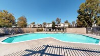Located at 3030 W Acacia Ave in Hemet, California, this outdoor community amenity space centers around a generously sized freeform swimming pool with crystal-clear blue water, ideal for recreation and relaxation under the abundant Southern California sunshine. The pool is framed by a wide concrete pool deck, providing ample space for lounging, and is enclosed by a combination of wrought iron fencing and block walls that ensure both security and privacy.

Multiple seating and relaxation zones are thoughtfully arranged throughout the area. A wooden deck extends from a covered pergola structure, offering a shaded retreat furnished with sling-style lounge chairs and a dining table — perfect for gathering out of direct sun. Additional sling loungers are positioned poolside along the concrete deck, allowing residents to enjoy open-air sunbathing.

The surrounding landscape features mature deciduous trees that provide natural shade and a backdrop of greenery, complementing the well-maintained lawn areas visible adjacent to the pool. The community's residential buildings — characterized by their neutral-toned, gabled-roof architecture — are visible in the background, reinforcing the cohesive neighborhood setting. The space benefits from abundant natural light and open sky views, creating a bright, welcoming atmosphere suited for both leisurely afternoons and active outdoor enjoyment.