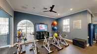Located at 2300 Kathryn Ln in Plano, Texas, this fitness center offers a comprehensive workout facility spread across a single, generously sized floor. The space is divided into two distinct functional zones seamlessly connected by a glass partition door, creating an organized and purposeful layout.

The main workout area is bright and open, lined with floor-to-ceiling windows that flood the space with natural light and offer views of the surrounding outdoor landscape. Ceiling fans and recessed lighting ensure a comfortable environment at all times. Large wall mirrors extend the visual depth of the room and support proper form during exercise. The area is well-stocked with a variety of strength-training machines, including cable systems and multi-use weight equipment, as well as cardio machines such as treadmills, ellipticals, and stair climbers. Wall-mounted televisions provide entertainment during workouts.

A dedicated cycling studio is separated by a sleek glass wall and door, featuring multiple stationary spin bikes arranged in front of a large flat-screen television. Colorful stability and exercise balls are stored on a wall-mounted rack, adding versatility for functional training. Crown molding, blue accent walls, and rubber flooring throughout lend the space a polished yet practical aesthetic, making this fitness center a well-rounded and inviting facility for all types of training.