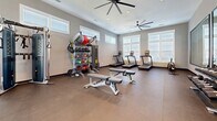 This fitness center offers a comprehensive workout environment designed to accommodate a wide range of exercise needs. The space is finished with durable rubber flooring throughout and features neutral gray walls that create a clean, focused atmosphere. Large windows with blinds line multiple walls, flooding the room with natural light, while oversized ceiling fans provide consistent airflow across the open floor plan.

The cardio zone is well-stocked with multiple treadmills, elliptical trainers, a stationary upright bike, and a stair climber, all positioned to take advantage of the natural light from surrounding windows. A dedicated strength and free weight area includes a tiered dumbbell rack paired with a large wall-mounted mirror, allowing users to monitor their form during workouts. Adjustable Matrix weight benches are centrally positioned for versatile use.

Functional training is supported by a multi-rack storage system holding medicine balls, kettlebells, resistance bands, and slam balls, alongside a cable-based functional trainer. A wall-mounted television and a small storage cabinet provide added convenience. Recessed lighting and ceiling-mounted fans ensure the space remains comfortable and well-lit at all times.