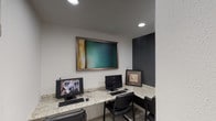 This single-story space functions as a professional business center, thoughtfully designed to support productivity and provide a clean, modern environment. The layout includes a dedicated computer workstation area and a larger open office, each with distinct design elements that contribute to a cohesive overall aesthetic.

The workstation area features an L-shaped granite countertop that wraps along the walls, providing ample surface space for multiple desktop computers and seating for several users simultaneously. Recessed lighting illuminates the workspace, while a large framed abstract artwork in teal and earth tones anchors one wall. A contrasting dark gray accent wall adds visual depth to the area, and decorative framed artwork provides additional character.

The main office area is bright and open, benefiting from a generous row of large windows along one wall that allow abundant natural light to flow throughout the space. Multiple workstations with desktop computers are arranged to take advantage of the exterior views. Light wood-style flooring runs through this area, complementing the crisp white walls and recessed ceiling lighting. A hallway connects the two areas, maintaining a smooth and functional flow throughout the space.