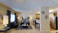 This single-floor residence presents an open-concept layout that connects the living area to the kitchen, creating a spacious and cohesive environment. The living room is anchored by plush carpeting and features a comfortable seating arrangement centered around a contemporary coffee table and a bold geometric area rug in brown and gray tones. A decorative wall display of circular mirrors serves as a striking focal point, complementing the neutral beige walls. Track lighting overhead provides adjustable illumination, while floor lamps add warm, ambient light. Natural light filters in through a window dressed with dark curtains. The kitchen is partially open to the living space, separated by a raised bar counter with seating for two, ideal for casual dining or entertaining. Warm wood cabinetry is visible in the kitchen area, adding a natural contrast to the light-toned countertops. Decorative wall accents, including a starburst piece, add personality to the space. The overall ambiance is modern and welcoming, blending functional design with curated décor throughout.