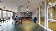 This expansive single-floor amenity lounge combines industrial design with a lively, social atmosphere. Exposed brick walls in muted gray and earthy tones define the space throughout, providing a raw, urban aesthetic that anchors the room's eclectic character. Large floor-to-ceiling windows along one wall flood the interior with abundant natural light, offering views of the surrounding landscape and creating an open, airy feel despite the room's depth and scale.

The flooring transitions between rich wood-toned planks and a textured teal-and-gold patterned carpet, visually delineating distinct activity zones. The social seating area features a mix of round tables paired with industrial-style metal bar stools and standard dining chairs, accommodating both casual gatherings and group seating. A plush lounge nook with a built-in bench and throw pillows provides a more relaxed setting near the windows.

The game zone is well-appointed, housing a shuffleboard table, a pool table, and a foosball table, all set against the dramatic brick backdrop. Overhead, a cascading cluster of amber and clear glass pendant lights adds warmth and visual interest to this corner. Accent seating—including teal leather chairs and bold blue velvet swivel chairs—creates intimate conversation spots throughout. Colorful billiard-themed artwork punctuates the walls, reinforcing the playful and social spirit of the space.