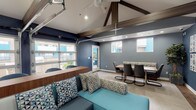 Located at 145 W Broadway in Long Beach, California, this single-floor community lounge space delivers a modern, urban aesthetic defined by bold navy blue walls, vaulted ceilings with exposed dark wood beams, and light wood-style flooring throughout. The architectural design draws the eye upward with dramatic ceiling trusses and a ceiling fan, creating an open and airy atmosphere despite the intimate scale of the room.

The lounge area is anchored by a teal sectional sofa accented with geometric-patterned throw pillows, complemented by a contemporary white accent table. A wall-mounted flat-screen television and curated large-format artwork panels add visual interest and a creative, gallery-like feel to the main seating zone. Adjacent to the lounge, a dedicated dining area features a solid wood table with modern cantilever chairs and a built-in banquette with patterned upholstery, offering a comfortable setting for group gatherings.

High windows along the walls invite natural light while providing glimpses of the surrounding outdoor courtyard. A full-panel glass garage-style door opens the space directly to the exterior, seamlessly blending indoor and outdoor living. A glass-paned entry door further reinforces the connection to the outdoor terrace area. Additional high-top tables and chairs are positioned near the windows, offering flexible seating options. The overall ambiance is vibrant, contemporary, and well-suited for social and community-oriented use.