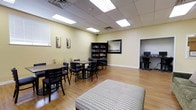 Located at 310 N Broad St in Carneys Point, New Jersey, this single-floor facility offers a thoughtfully arranged communal environment designed for comfort and functionality. The main gathering area features warm yellow-toned walls, a drop-panel ceiling with recessed fluorescent lighting, and durable hardwood-style flooring that runs throughout the space, creating a bright and inviting atmosphere.

The room is furnished with multiple dining-style tables and black ladder-back chairs, providing ample seating for group gatherings or casual meals. A plush, upholstered ottoman adds a comfortable lounge element to the space. Along one wall, a dark wood bookcase holds a curated collection of books, making it an ideal spot for reading and quiet reflection. Framed artwork decorates the walls, contributing a homey, residential feel to the interior.

An adjacent alcove, visible through an open doorway, houses a computer workstation, offering a dedicated area for residents or visitors to access digital resources. The overall layout is open and easy to navigate, with natural light filtering in through window treatments on one side. The space strikes a balance between practicality and comfort, making it well-suited as a community lounge, activity room, or resource center for the building's occupants.