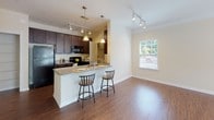 This single-floor residence combines practical design with comfortable living across several well-appointed spaces. The open-concept kitchen serves as a central hub, showcasing dark espresso cabinetry, granite countertops, and black appliances including a refrigerator and range. A peninsula with bar seating accommodates two stools beneath elegant pendant lighting, while track lighting above adds additional illumination. The kitchen flows naturally into the living and dining areas, which feature warm hardwood-style flooring throughout. Crown molding, a ceiling fan, and French doors leading to a balcony or patio distinguish the living area, allowing natural light to filter in freely. A built-in shelving niche provides a functional and decorative accent along one wall. The bedroom is carpeted and benefits from a generously sized window with blinds offering outdoor views. The bathroom features a dark-cabineted vanity with granite countertop and a shower-tub combination with tiled surround. Completing the layout, an in-unit laundry area houses a full-sized washer with overhead wire shelving, adding everyday convenience to this thoughtfully designed residence.